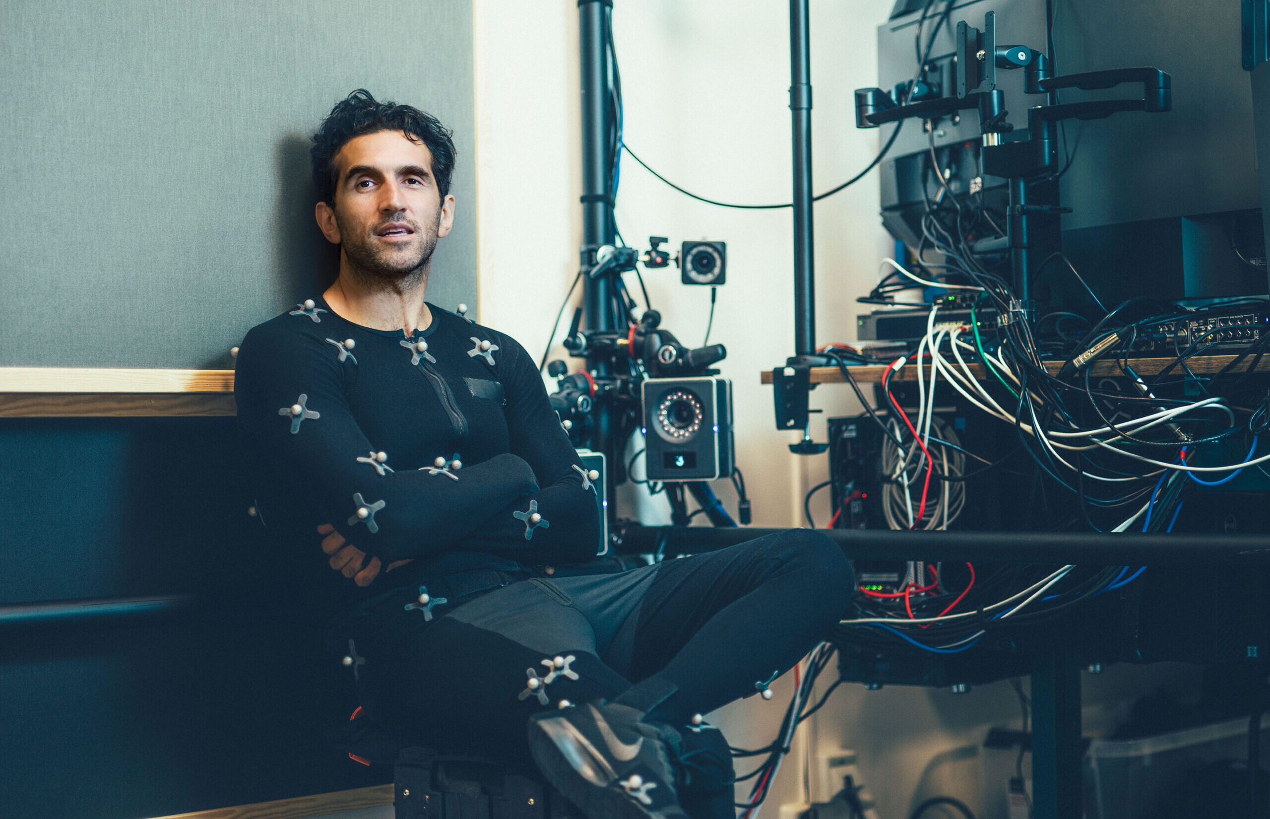 Hazelights oprichter Josef Fares leunt achterover in zijn zwarte mocap-pak met witte bolletjes erop. Hij kijkt naar de camera. Achter hem zijn draden en machines te zien.
