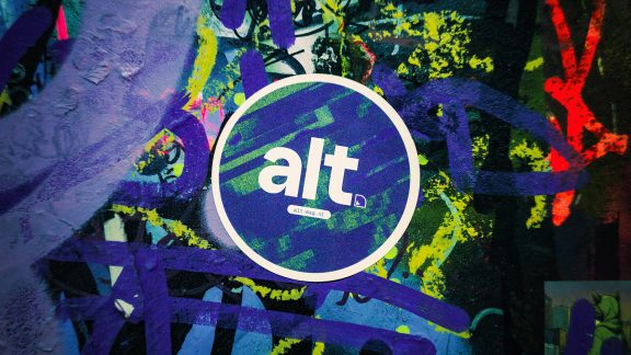 Logo van alt. op een sticker die op een muur vol grafiti is geplakt.