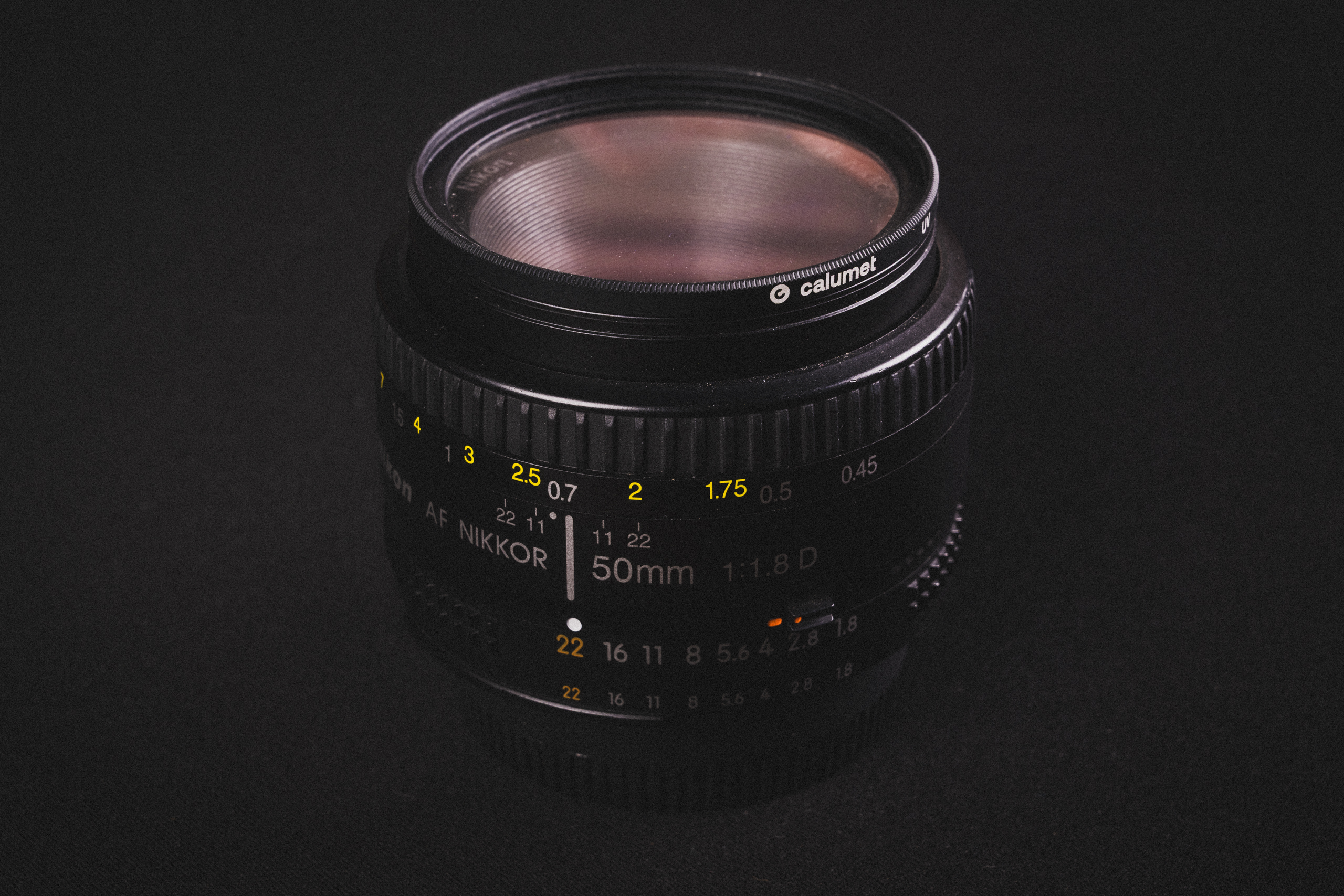Nikkor 50mm f/1.8-AF-D op een zwarte achtergrond. De lens staat rechtop, zonder lenskap (maar met UV-filter).