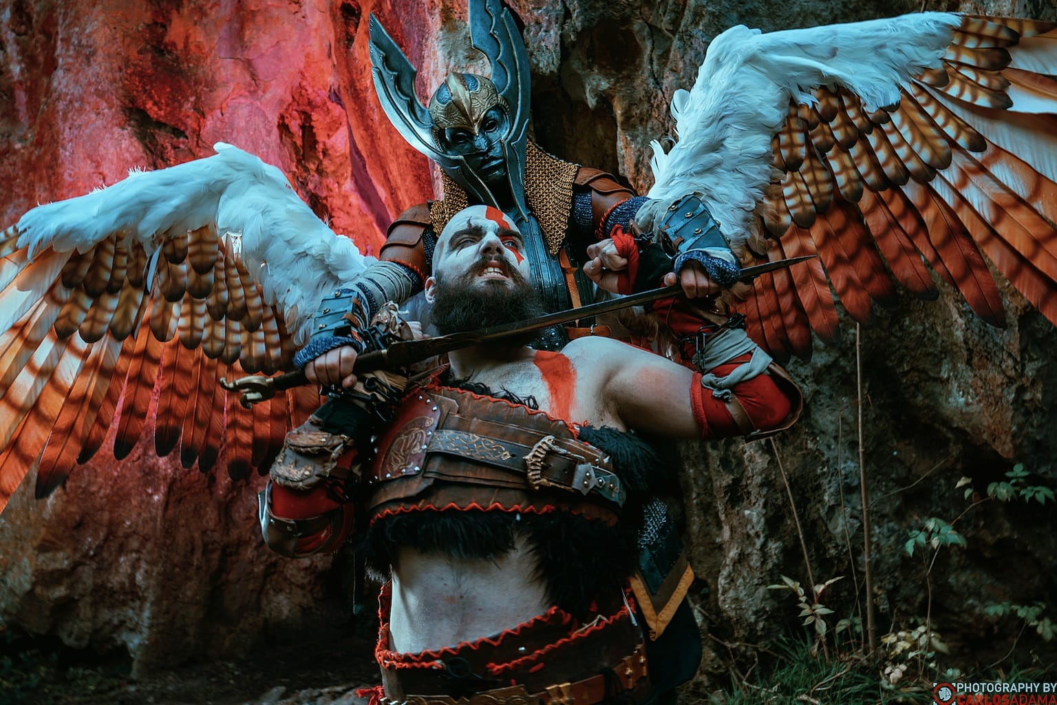 Kratos, een brede man met een baard en rode warpaint, wordt van achteren met een zwaard aangevallen door Freya, een gevleugeld personage met een helm die ook een masker is. Op de achtergrond zijn rood belichte rotsen te zien.