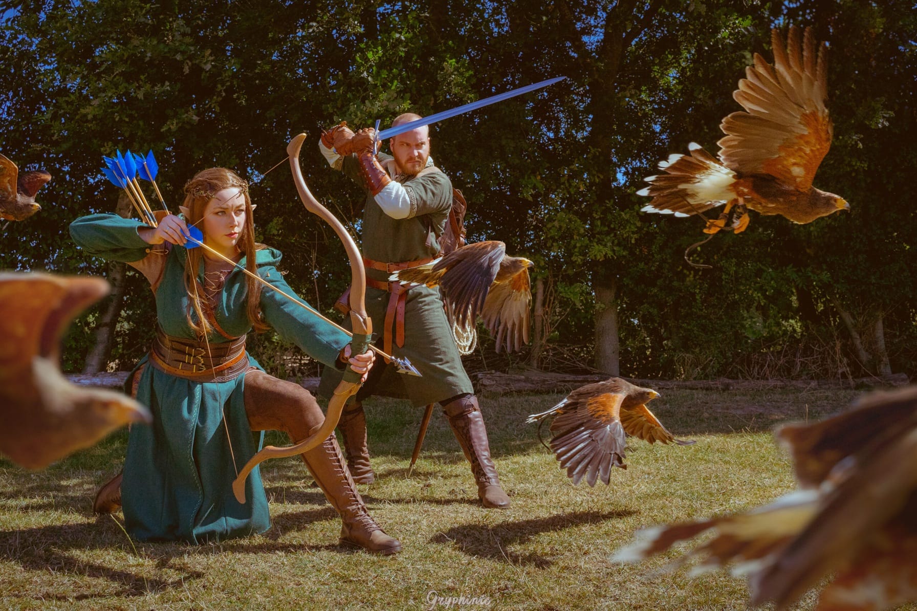 Twee cosplayers in fantasy-outfits staan klaar in actieposes. Een heeft een zwarad vast, de ander pijl en boog. Zij kijken ernstig naar iets buiten beeld. Op de voorgrond vliegen meerdere roofvogels.